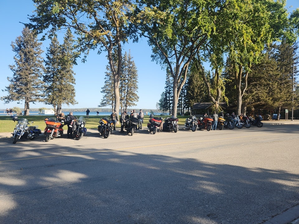 Chapter bikes lined up in a lakeside park with tall pines and blue water in the background