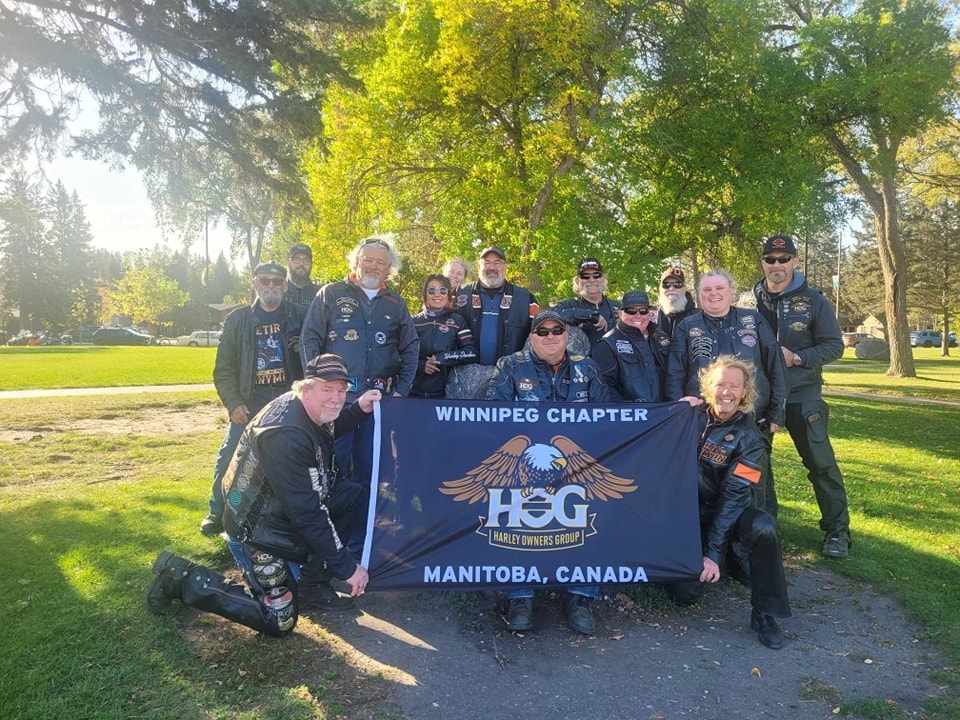 Group of riders holding the Winnipeg Chapter HOG Manitoba Canada banner in a sunny park