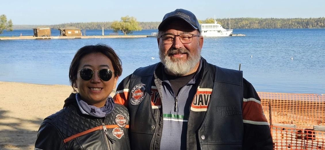 Two chapter members in Harley-Davidson vests smiling at a sandy lakeside beach with a tour boat in the background