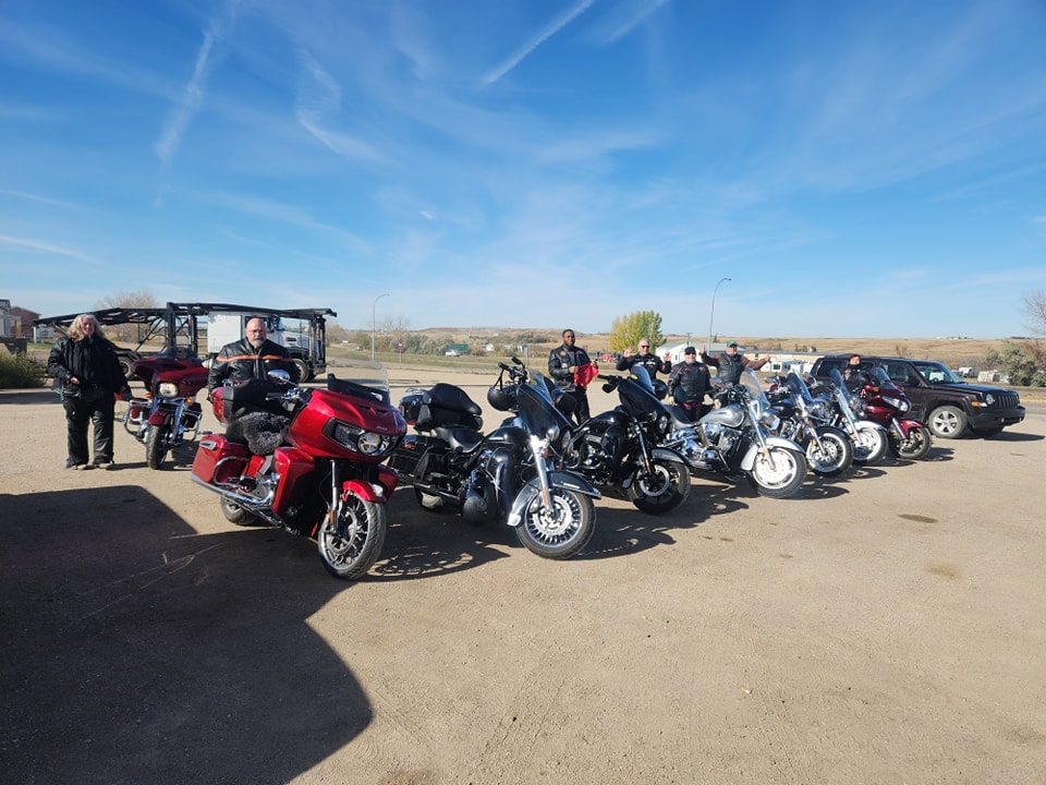 Group of chapter riders and bikes lined up under a wide blue prairie sky at a ride stop