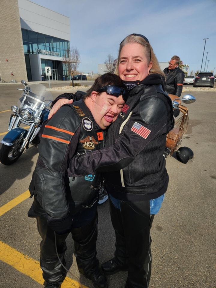 Two chapter members sharing a warm hug in a parking lot with bikes in the background