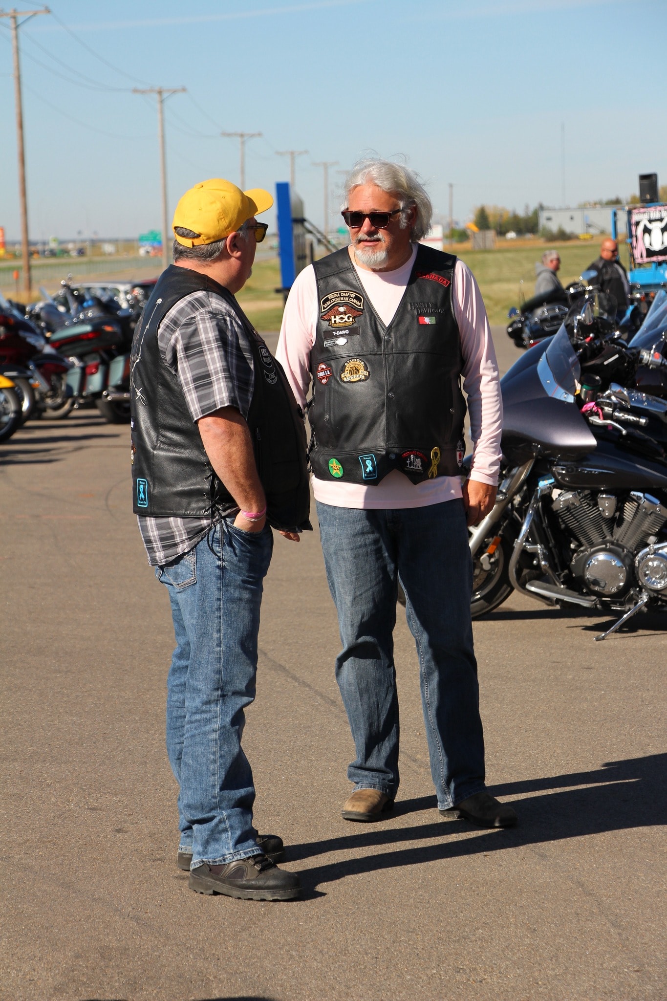 Two chapter members in HOG vests chatting in the parking lot surrounded by bikes at the 2024 Breast Cancer Awareness Ride