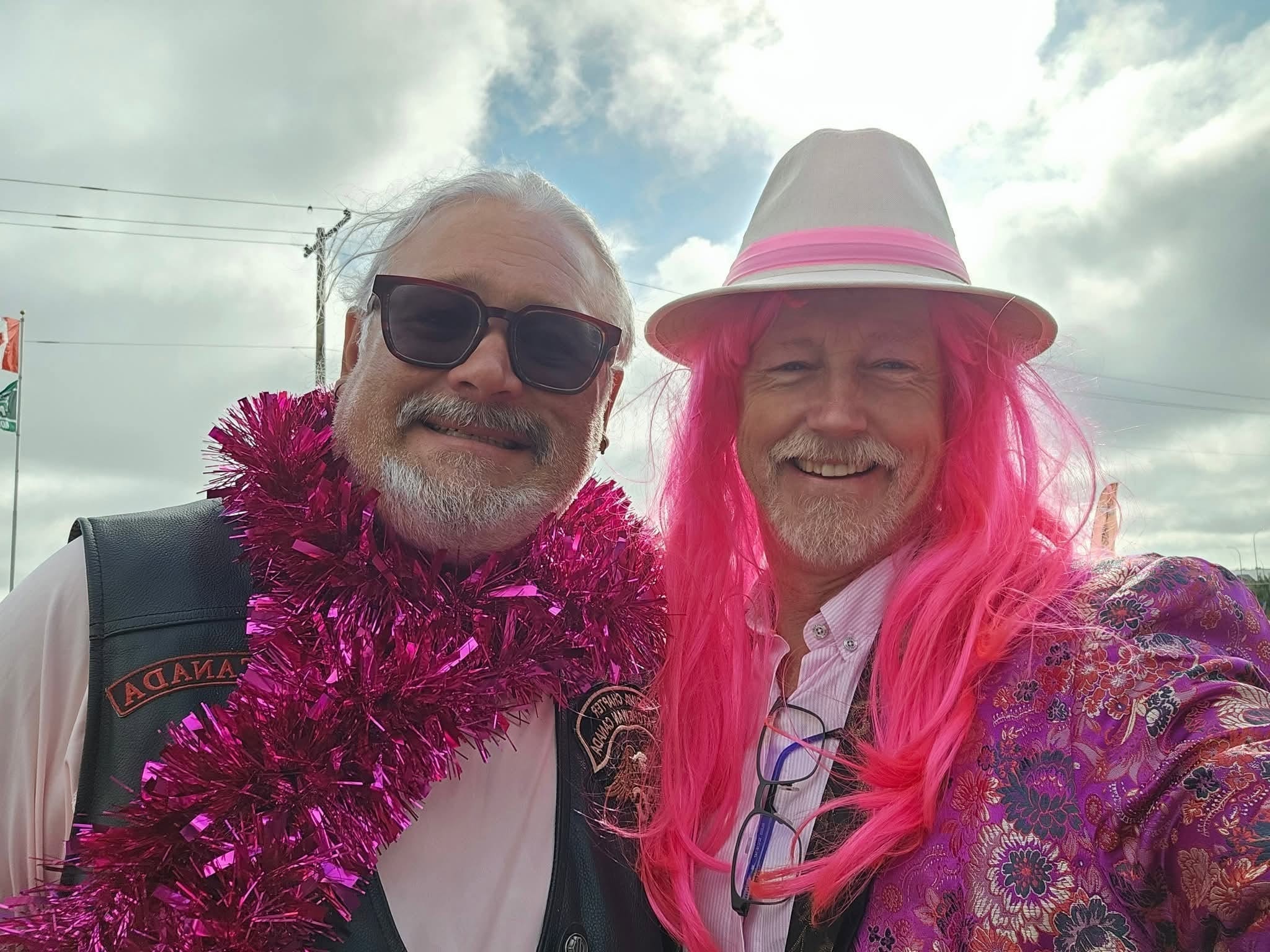 Two chapter members grinning at the 2025 Breast Cancer Awareness Ride — one in a HOG vest with a pink tinsel boa, one in a pink wig, white fedora and floral blazer