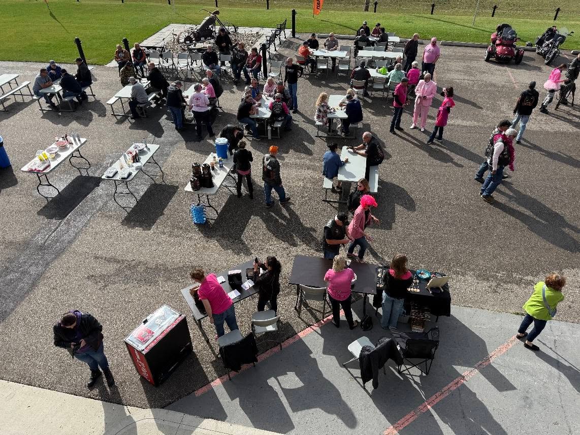Aerial view of the full 2025 Breast Cancer Awareness Ride event at Prairie Harley-Davidson, with dozens of riders at tables, vendor booths, and a pink-suited emcee visible in the crowd
