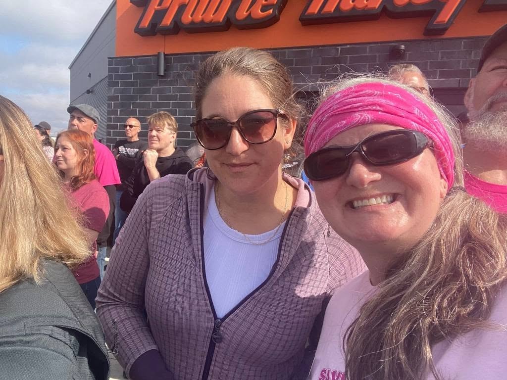 Two women smiling in front of the Prairie Harley-Davidson sign at the 2025 Breast Cancer Awareness Ride — one in a pink bandana headband and Save the Tatas shirt
