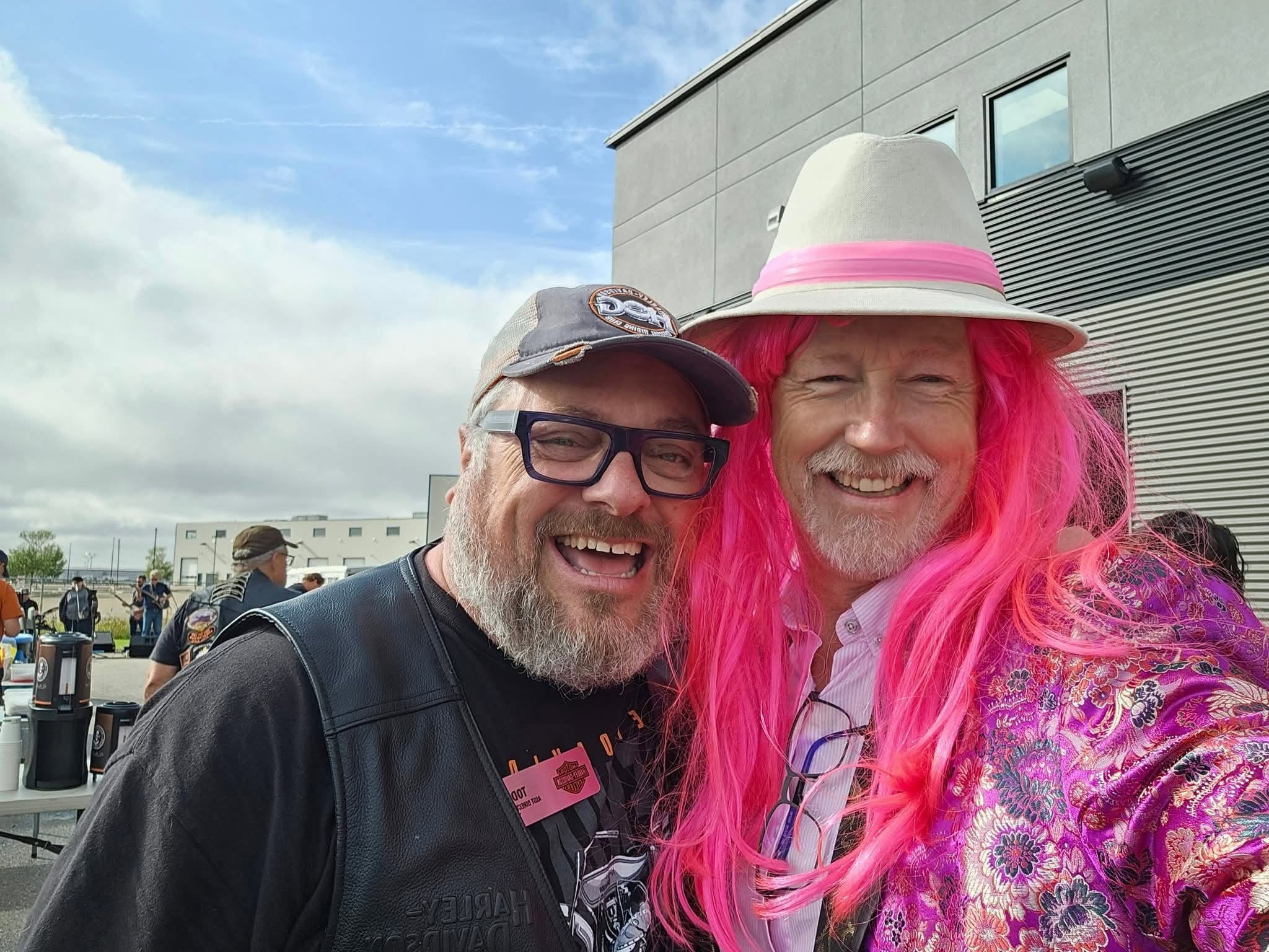 Two chapter members laughing together outside Prairie HD — one in a leather HOG vest, one in a bright pink wig and floral kimono at the 2025 Breast Cancer Awareness Ride