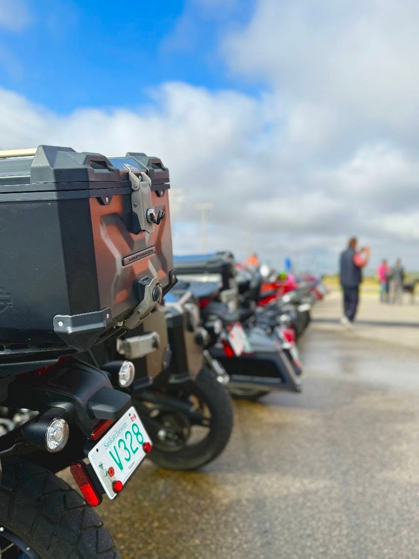 Close-up of a row of Harley-Davidson bikes lined up at the 2025 Breast Cancer Awareness Ride, with a Saskatchewan plate V328 in the foreground under a blue sky