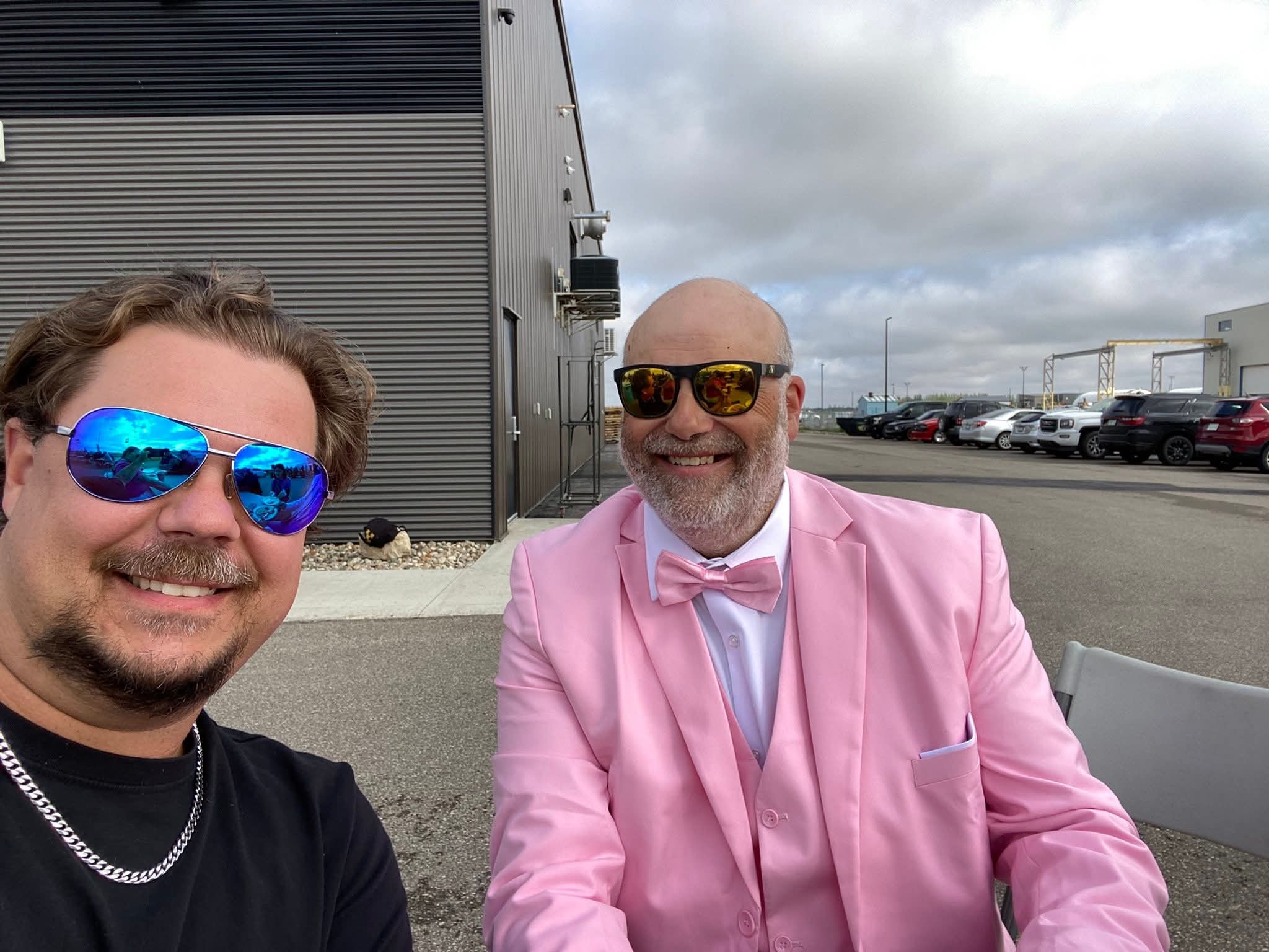 Two chapter members taking a selfie outside Prairie Harley-Davidson at the 2025 Breast Cancer Awareness Ride — one in a black t-shirt and blue aviators, one in a full pink three-piece suit and gold mirrored sunglasses