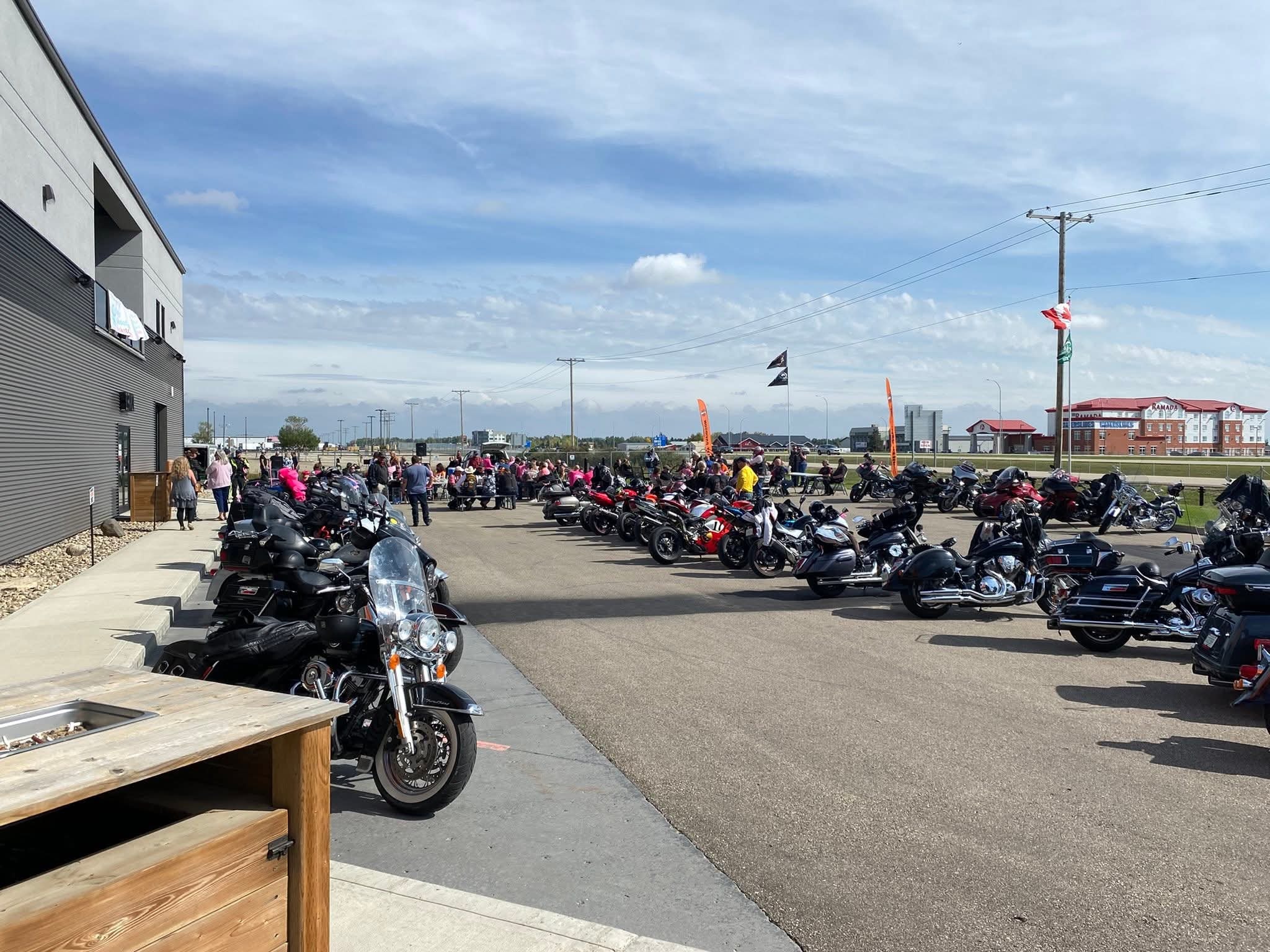 Wide shot of the 2025 Breast Cancer Awareness Ride at Prairie Harley-Davidson — dozens of bikes lined up in the parking lot, riders in pink milling about under a blue sky with orange event banners flying