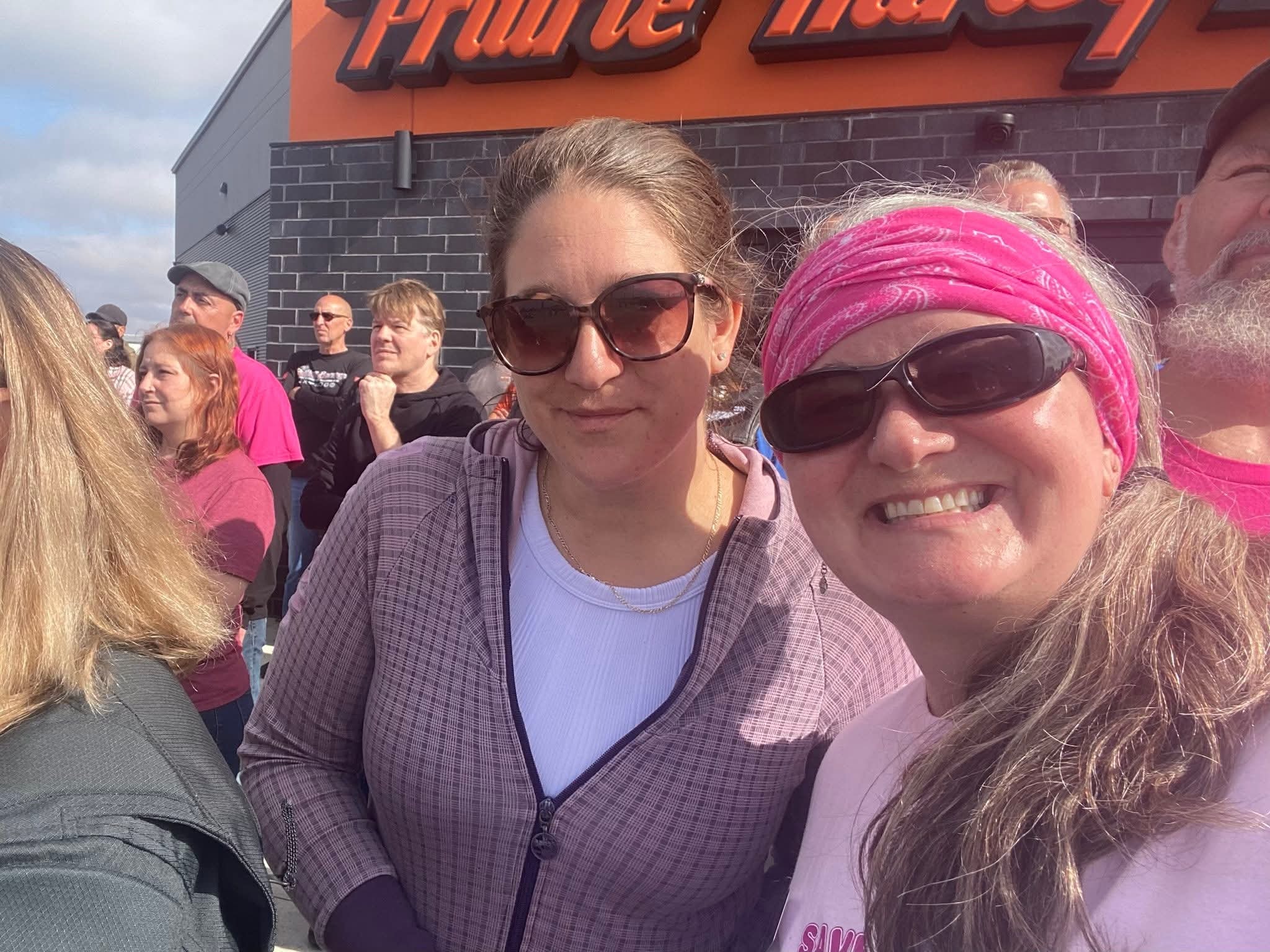 Two women smiling in front of the Prairie Harley-Davidson sign at the 2025 Breast Cancer Awareness Ride — one in a pink bandana headband and Save the Tatas shirt, crowd gathered behind them