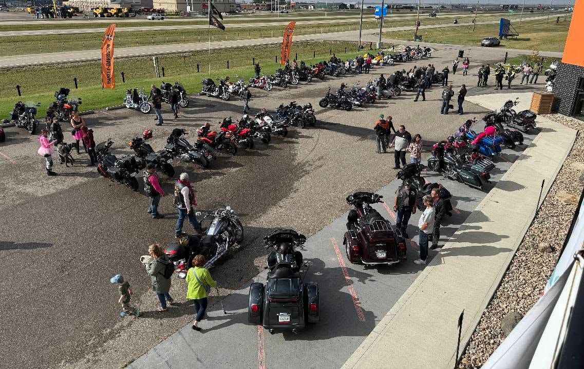 Three chapter members in the Prairie HD parking lot — one spreading arms wide, two making a heart shape with their hands — surrounded by bikes at the 2025 Breast Cancer Awareness Ride