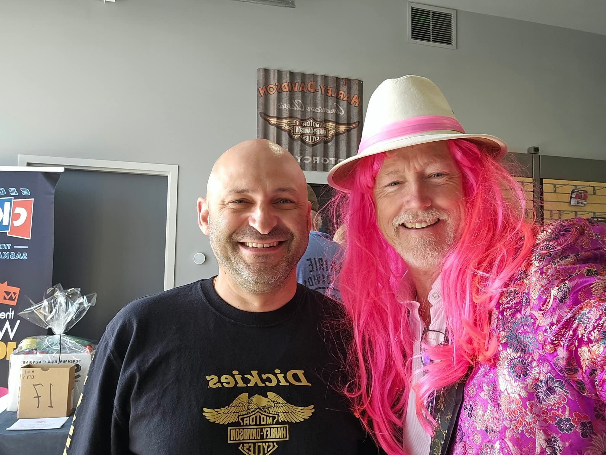 Two men smiling inside Prairie Harley-Davidson at the 2025 Breast Cancer Awareness Ride — one in a Dickies Harley-Davidson shirt beside a prize table, one in a bright pink wig, white fedora and floral kimono, a vintage HD tin sign on the wall behind them