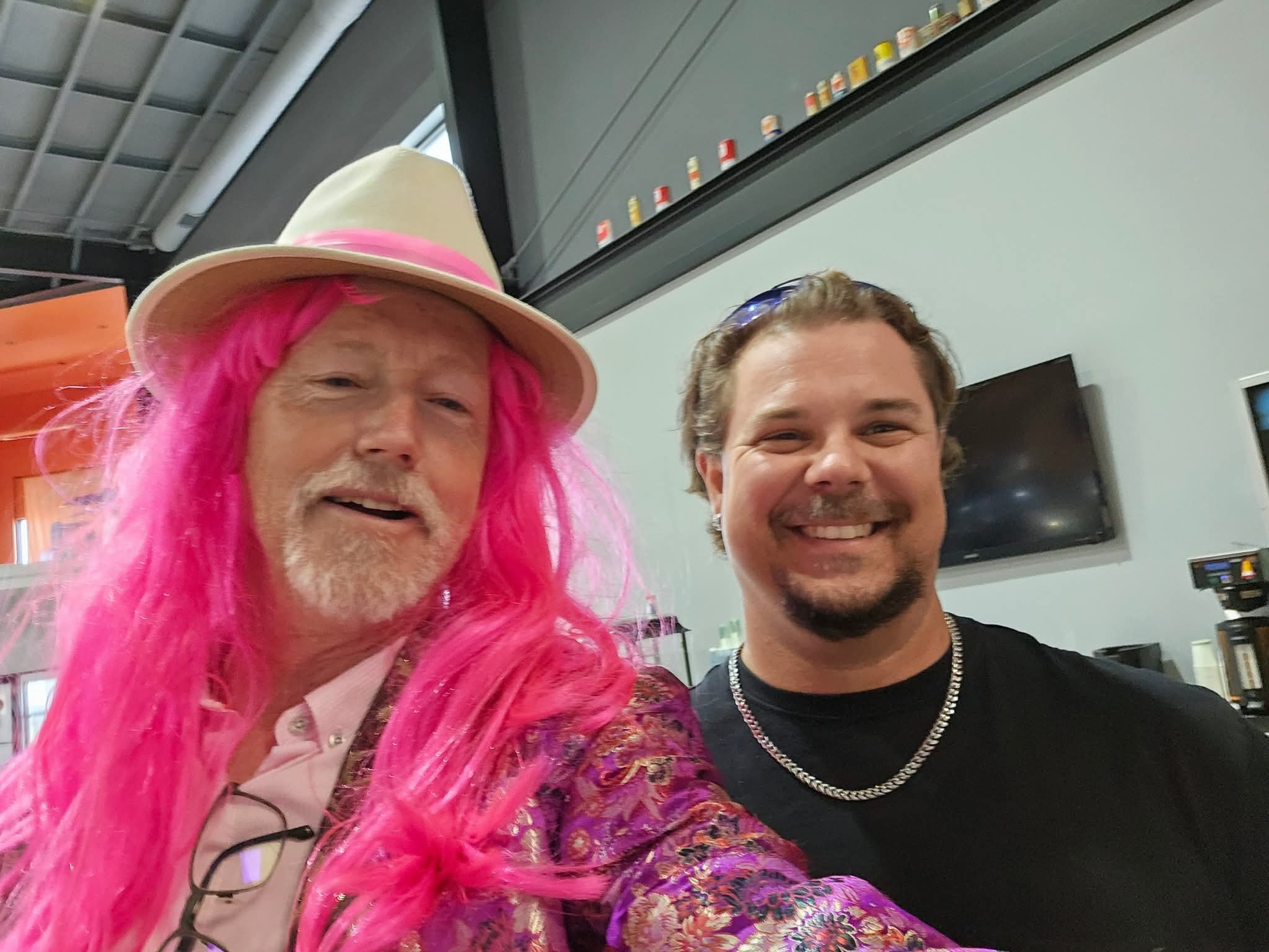 Two men taking a selfie inside Prairie Harley-Davidson at the 2025 Breast Cancer Awareness Ride — one in a bright pink wig, white fedora and floral kimono, one in a black t-shirt and chain necklace, vintage pop cans lining a shelf in the background