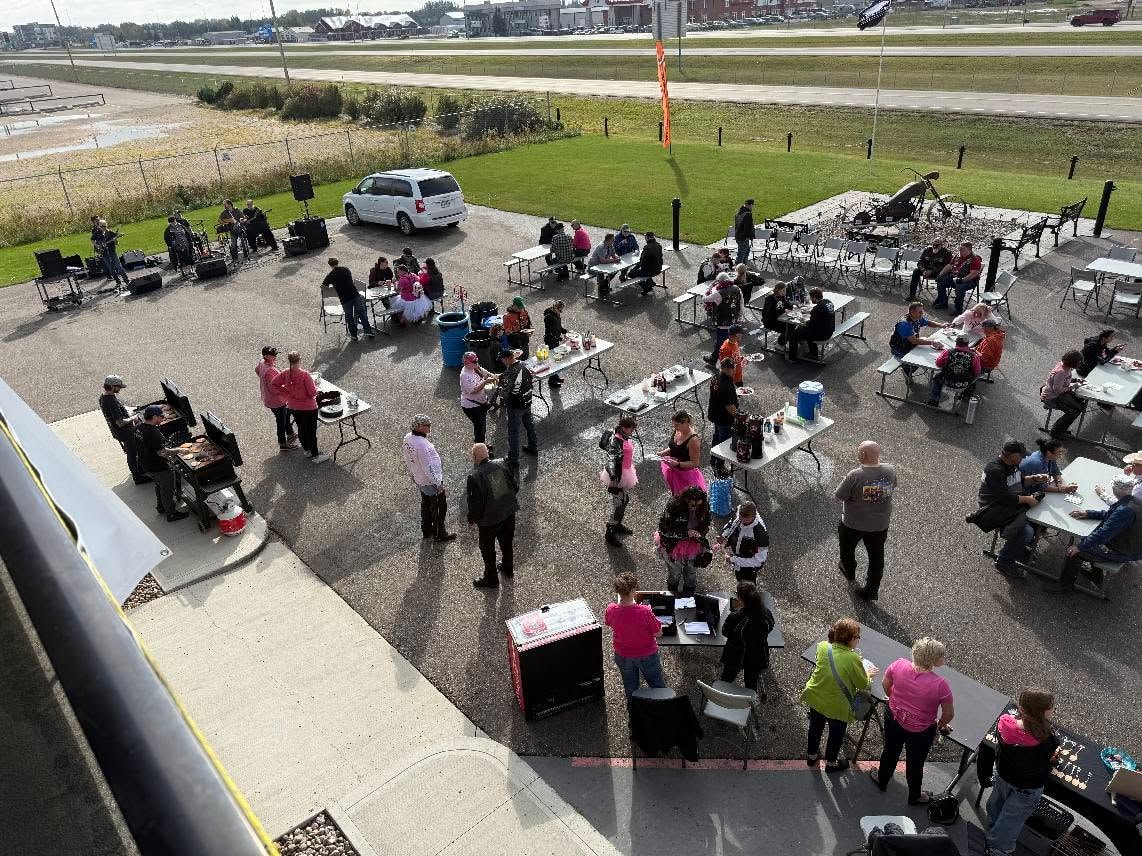 Aerial view of the 2025 Breast Cancer Awareness Ride breakfast at Prairie Harley-Davidson, with riders and volunteers spread across outdoor tables, a live band, and a grill station